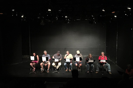 Several people sit on a dark stage performing a reading.